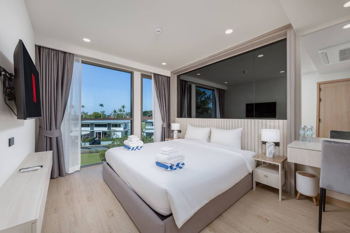 A modern hotel room with a large bed, a desk, a TV, and a balcony with a view of a building and greenery.