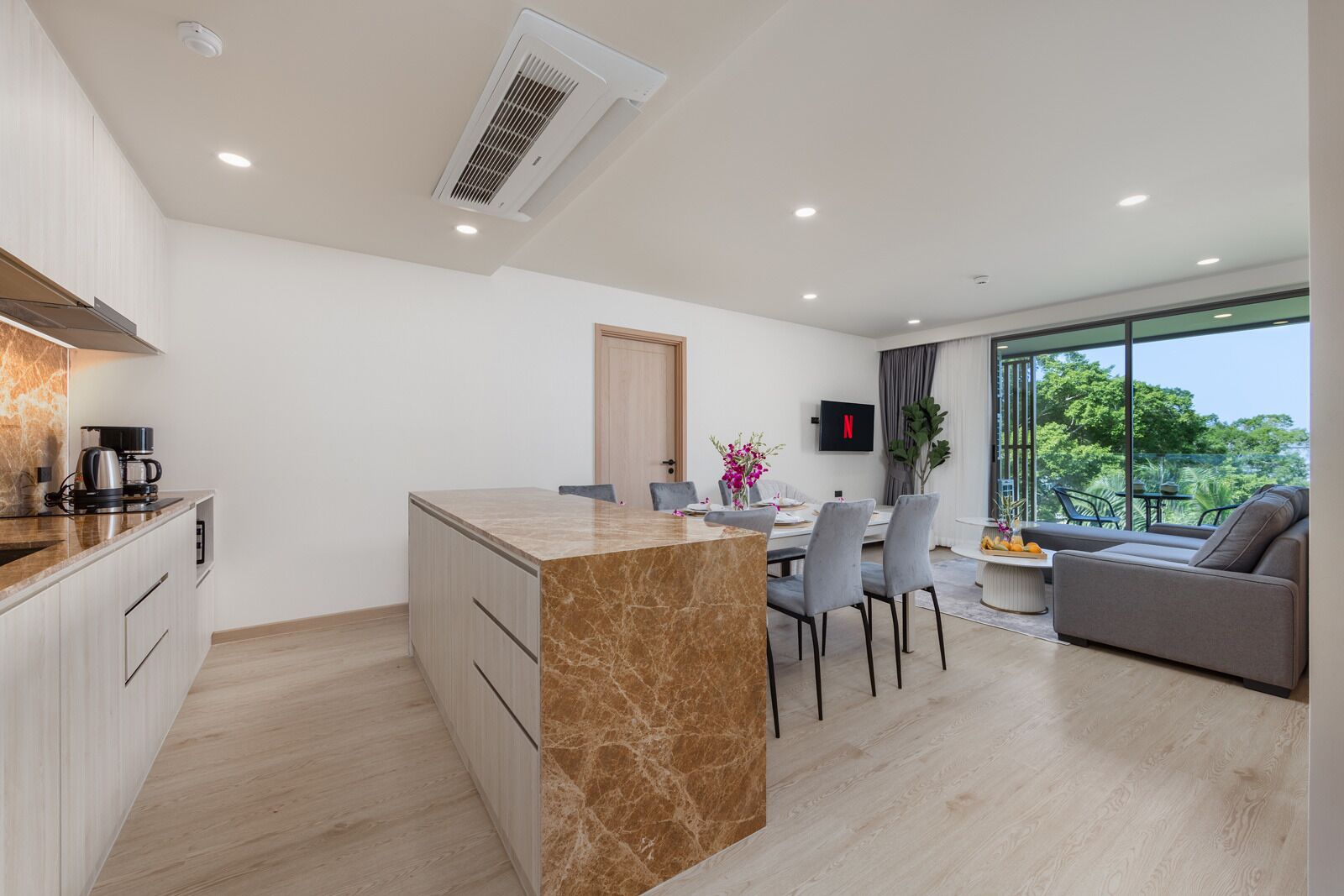 A modern apartment with a kitchen, dining area, and living room. It features a marble island, a flat-screen TV, and a balcony with a view of trees.