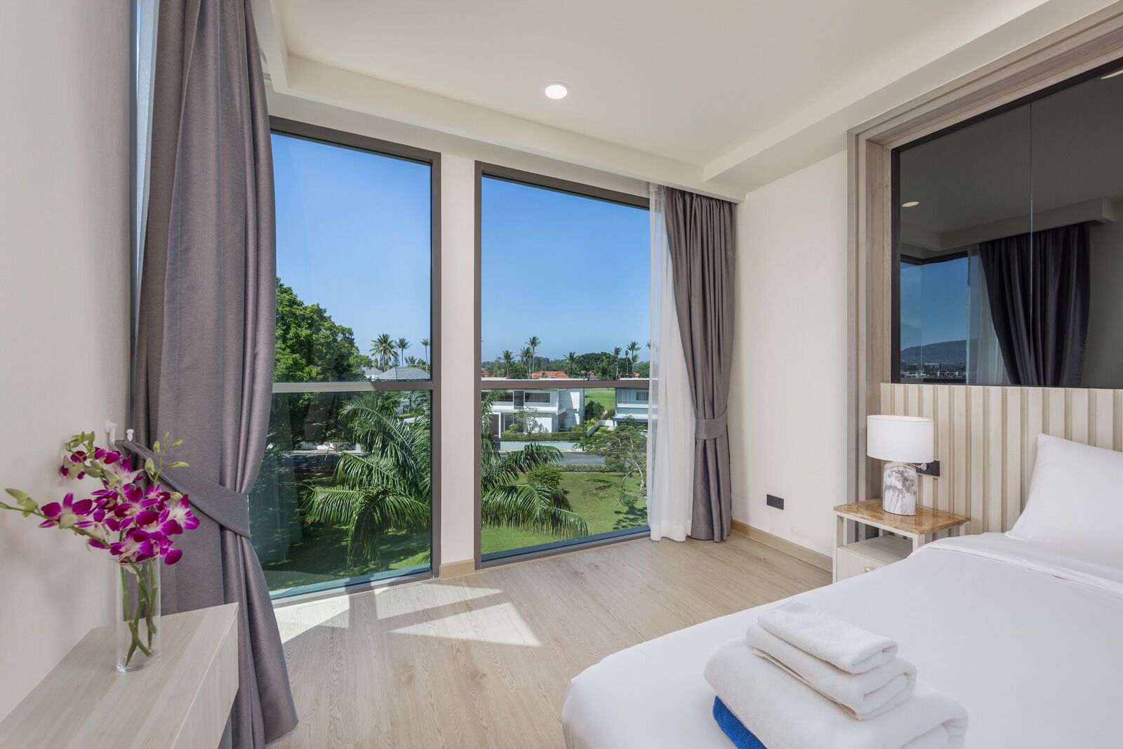 Una camera d'hotel moderna con un letto grande, un comodino, una lampada e vista su un paesaggio tropicale attraverso ampie finestre a tutta parete.