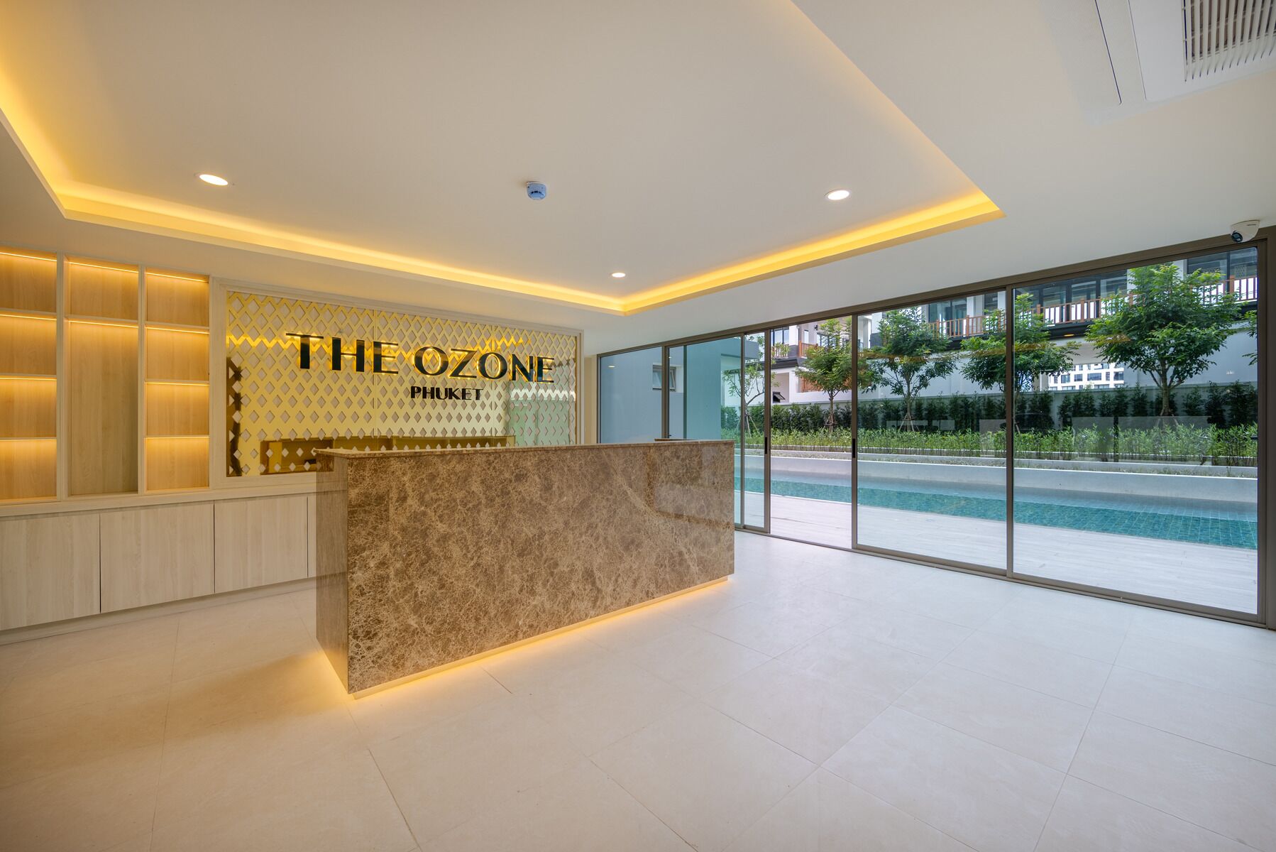 Un área de recepción con mostrador de mármol, una pared con la señalización 'THE OZONE PHUKET' y un amplio ventanal con vistas a una piscina y vegetación.