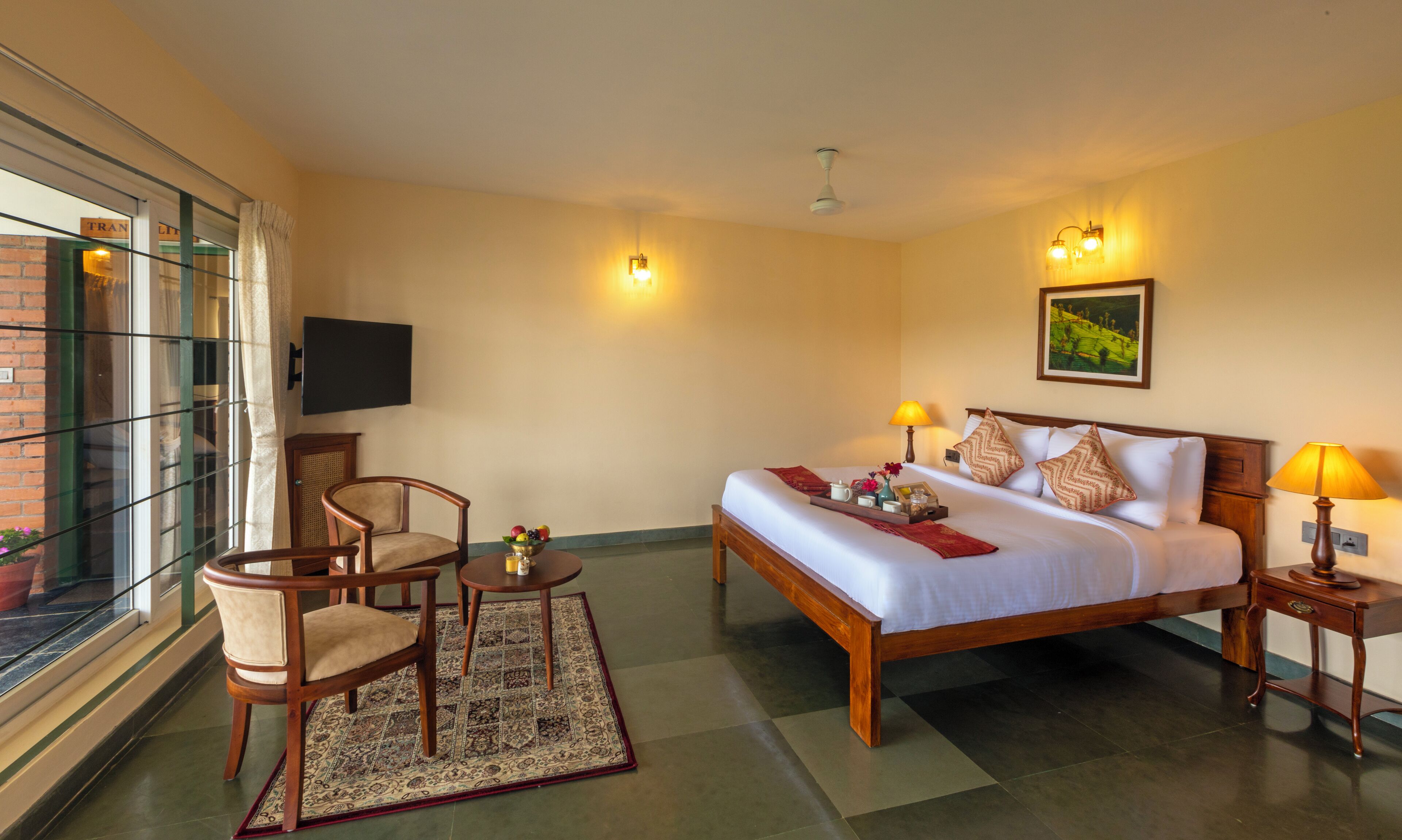 A hotel room with a bed, two bedside tables, a chair, a small table, a TV, and a window with curtains.