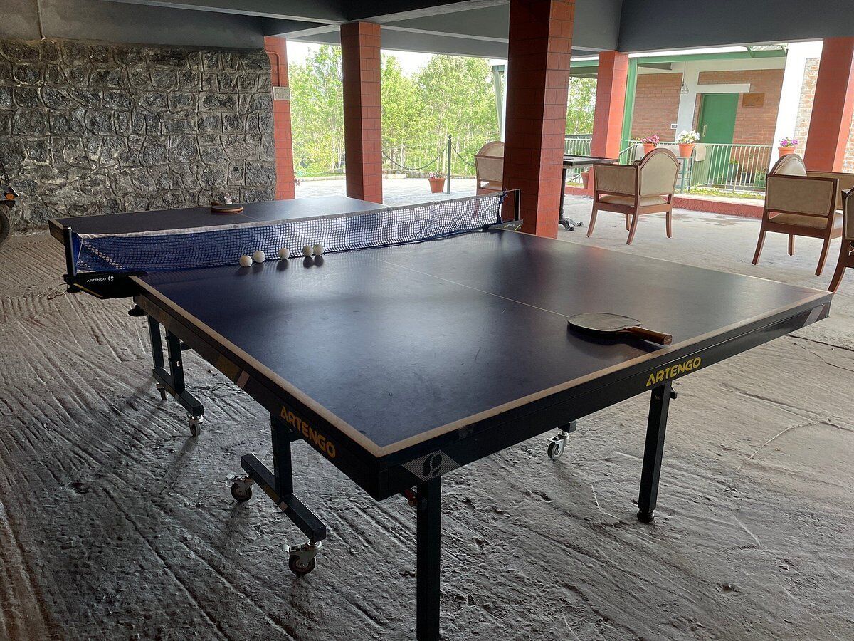 A ping pong table with balls and a paddle, set up on a wooden floor with a stone wall and chairs in the background.