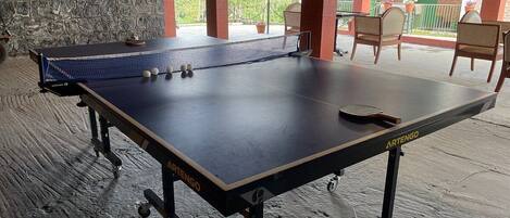 A ping pong table with balls and a paddle, set up on a wooden floor with a stone wall and chairs in the background.