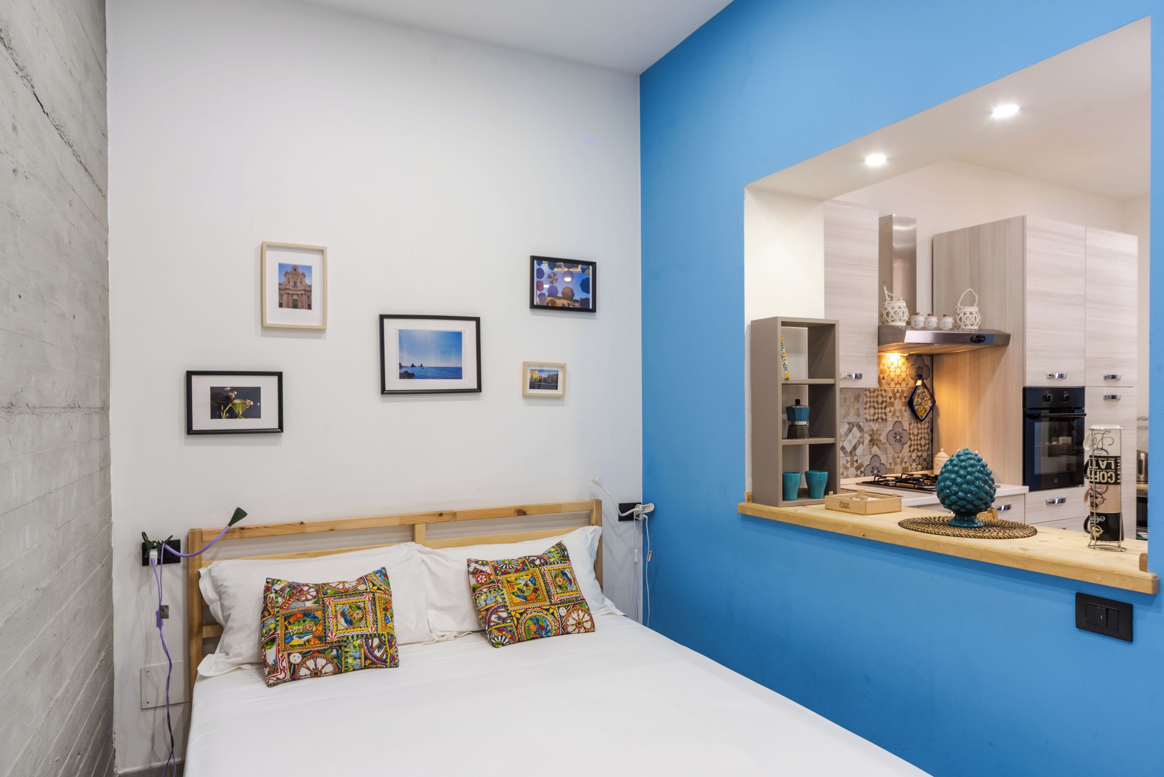 A bedroom with a bed, colorful pillows, a blue accent wall, and a kitchen visible through a mirror.