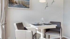 A dining area with a white table set for two, two chairs, a painting of a beach scene, and a modern light fixture.