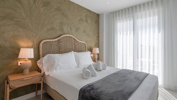 Ein ordentlich bezogenes Bett mit weißen Laken und einer dunklen Tagesdecke, begleitet von zwei Nachttischen mit Lampen, in einem Zimmer mit großem Fenster und leichten Vorhängen.