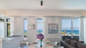 A modern dining area with a wooden table set for six, large windows offering a view of the sea, and a cozy seating area with a sofa.