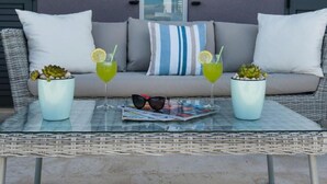 A wicker sofa with cushions, two potted plants, two drinks with lemon slices, and a pair of sunglasses on a glass table.