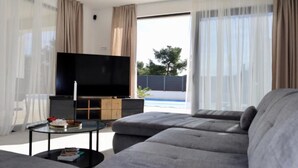 Una sala de estar moderna con un televisor de pantalla plana grande, sofás grises, una mesa de centro de vidrio y cortinas traslúcidas.