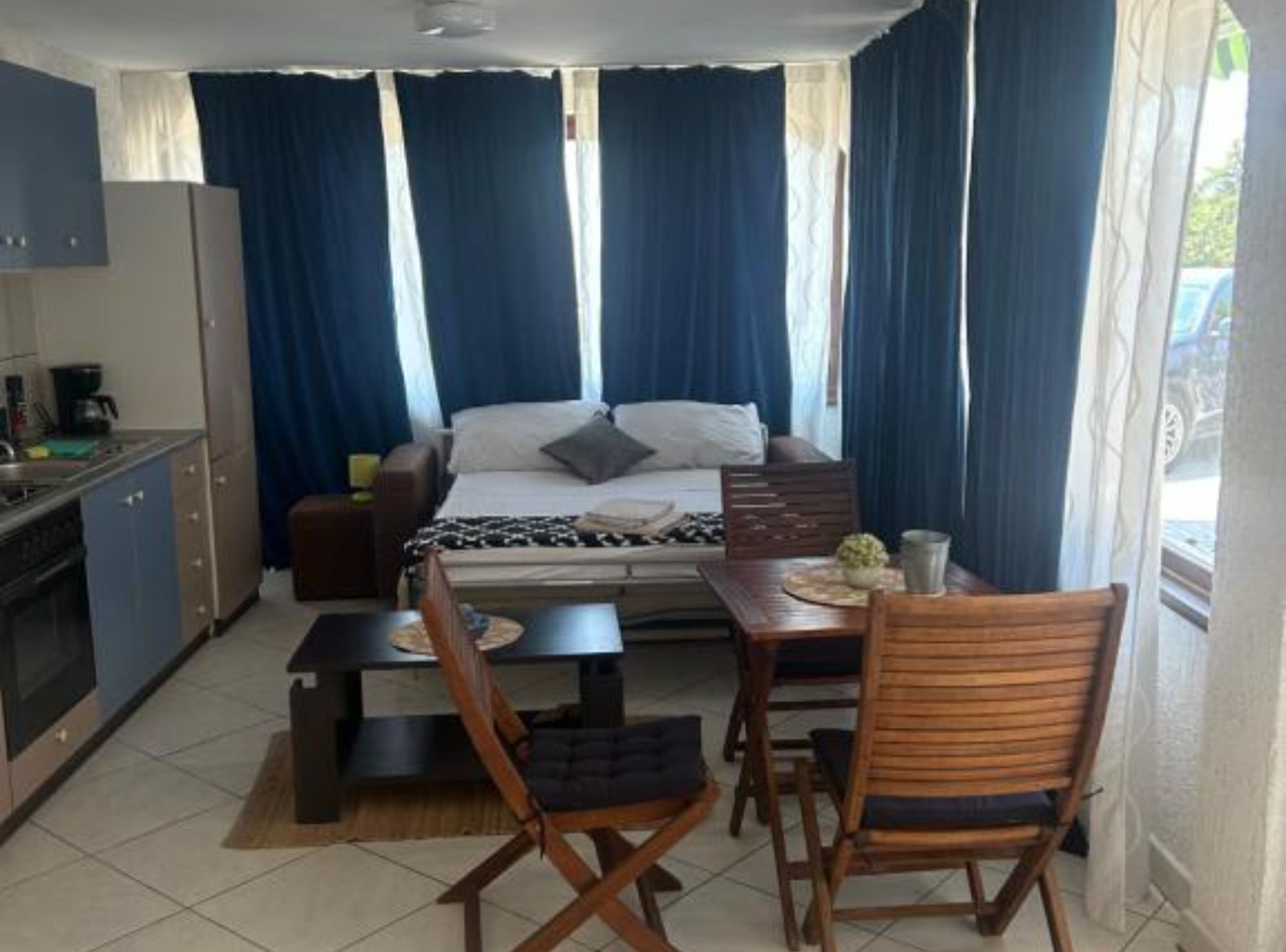 A compact room with a bed, a small kitchenette, a dining area with a table and chairs, and a window with curtains.