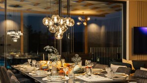 A modern dining area with a long table set for a meal, a chandelier, and a large window with a view. - Luxury Villa Lara with Heated Pool, Hot-Tub and Sauna (Zmijavci)