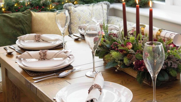Una mesa de comedor bien dispuesta con platos, vasos y velas, rodeada de follaje festivo y adornos navideños.