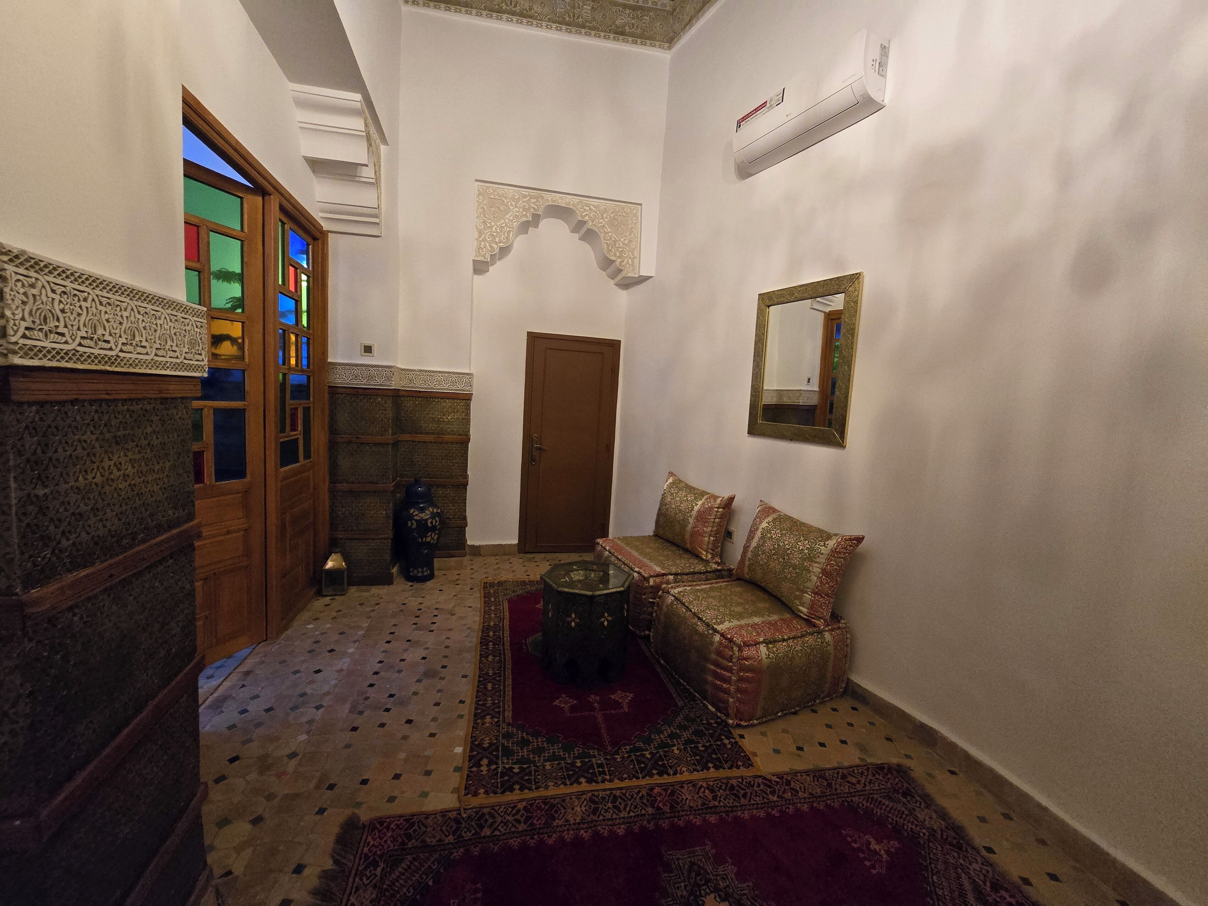 A narrow hallway with a wooden door, a small seating area with cushions, a patterned rug, and a wall-mounted mirror.