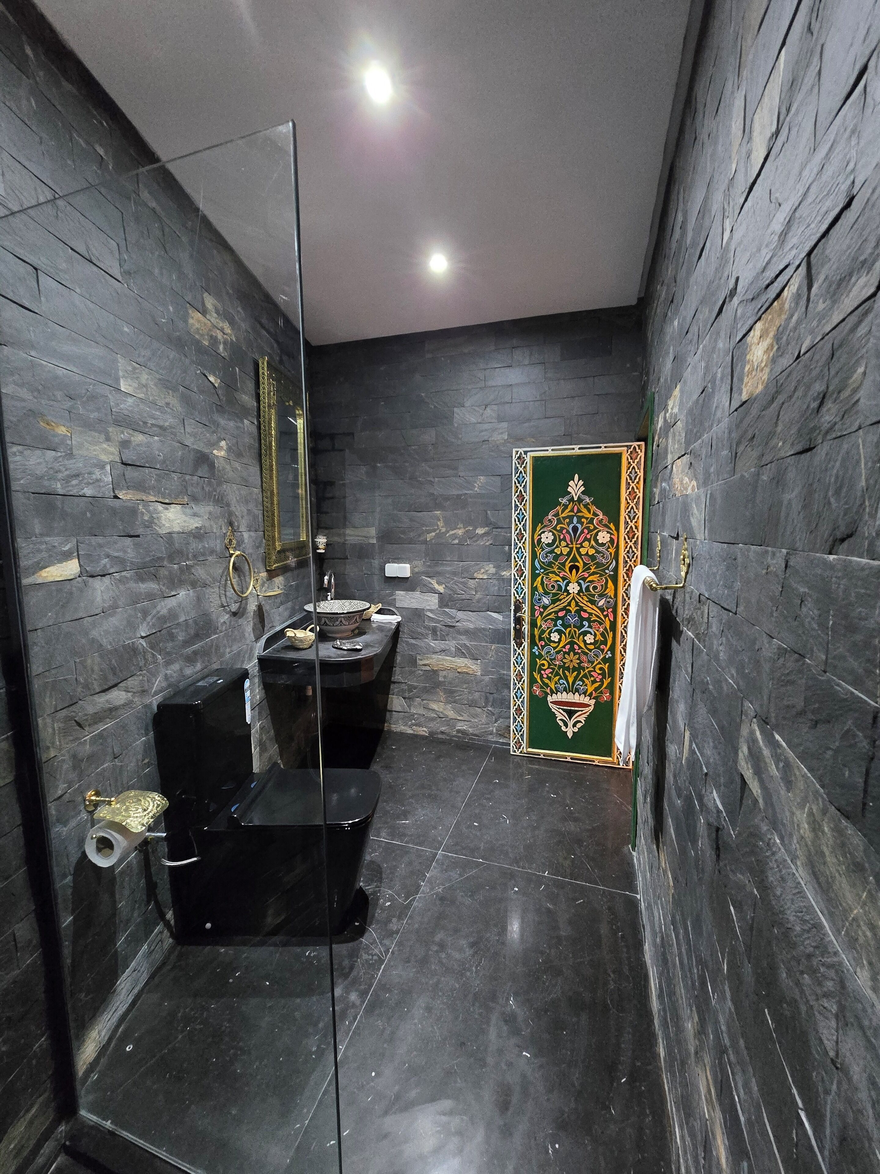 Um banheiro com piso de mármore preto, pia preta, vaso sanitário e um espelho. Há um painel decorativo na parede.