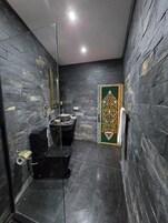Um banheiro com piso de mármore preto, pia preta, vaso sanitário e um espelho. Há um painel decorativo na parede.