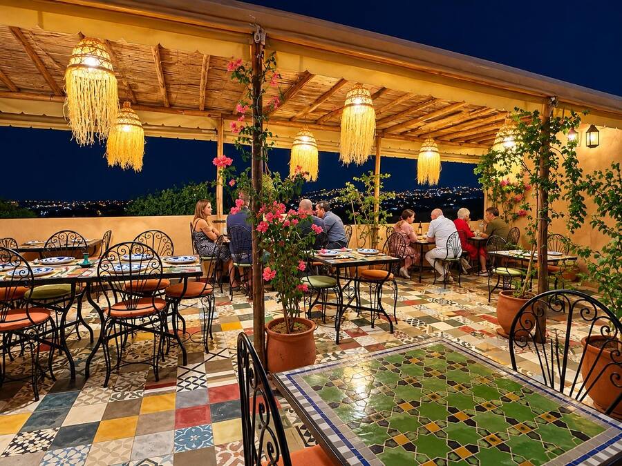 An outdoor dining area with tables and chairs, overhead lighting, and a view of the cityscape at night.