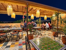 Un área de comedor al aire libre con mesas y sillas, iluminación colgante y vistas de la ciudad de noche.