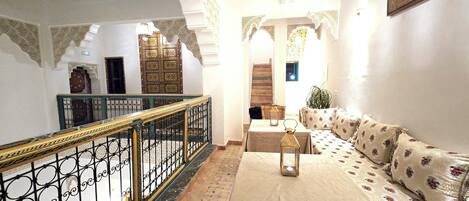 Quarto com teto alto, arcos ornamentados, escadaria, conjunto de sofás e mesa com um candeeiro.