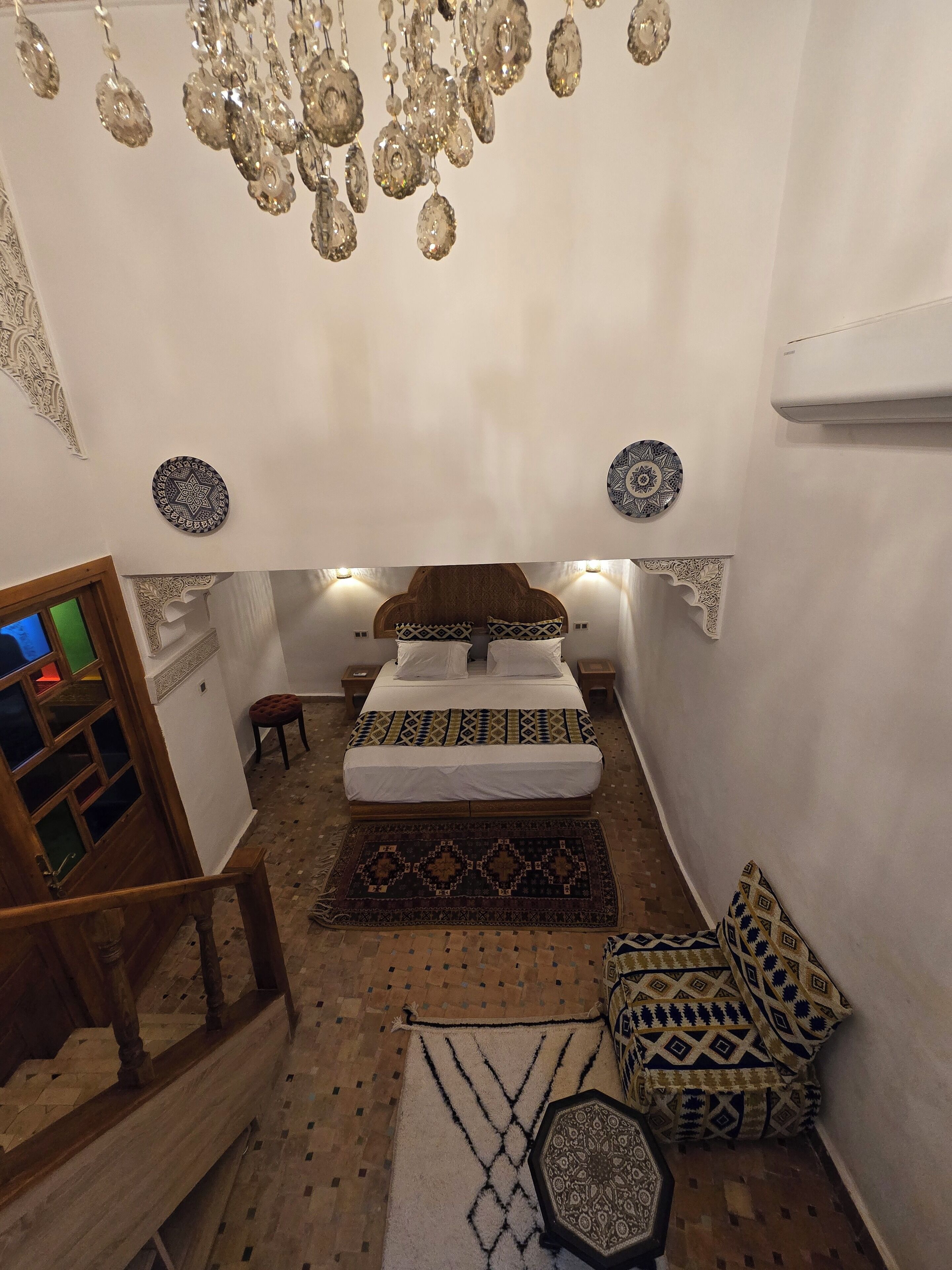 A bedroom with a bed, patterned pillows, a hanging chandelier, and a decorative rug.