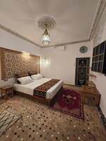 Quarto com uma cama grande, uma cabeceira decorativa, um candelabro, um tapete estampado e um banquinho de madeira.
