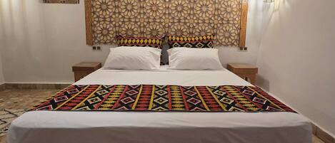 A bedroom with a large bed, a patterned headboard, a decorative wall hanging, and a small bedside table.