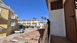 A residential street with a white car parked on the side, a staircase leading to a building, and palm trees in the background.