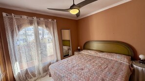 A bedroom with a bed, a ceiling fan, a nightstand, a mirror, and sheer curtains with a view of buildings outside.