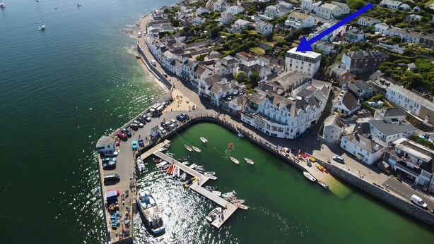Une vue aérienne d’une ville côtière avec un port de plaisance, des bâtiments blancs et une grande flèche bleue bien visible, indiquant un endroit précis.
