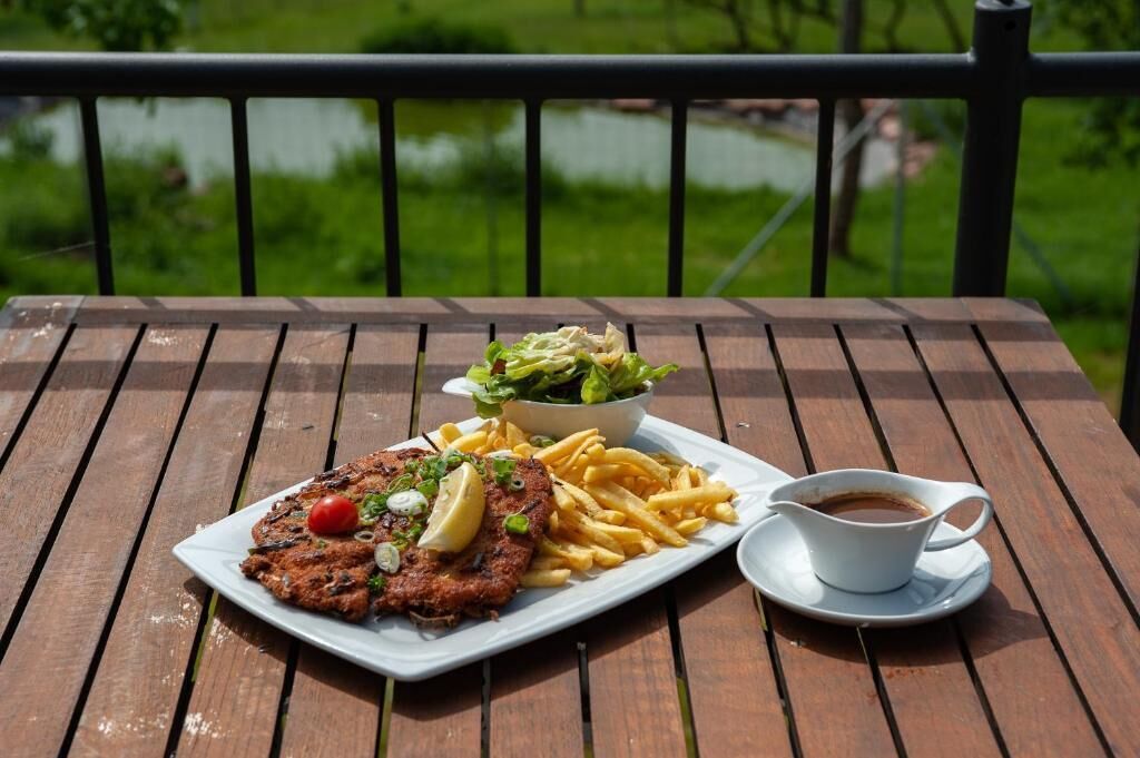 A meal consisting of a breaded cutlet, fries, a salad, and a cup of coffee on a wooden table with a view of greenery and a body of water in the background.
