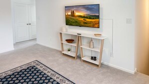 Ein Flur mit einer weißen Tür, einem an der Wand befestigten Fernseher, der eine Landschaft zeigt, einem weißen Konsolentisch mit Ablagen und einem gemusterten Teppich.