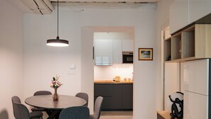 A modern kitchen with white cabinets, a microwave, and a toaster. There is a dining area with a round table and grey chairs. A vase with flowers is on the table.