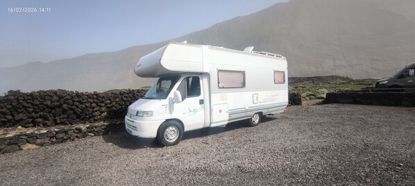 A white camper van parked on a gravel surface with a stone wall and mountains in the background.