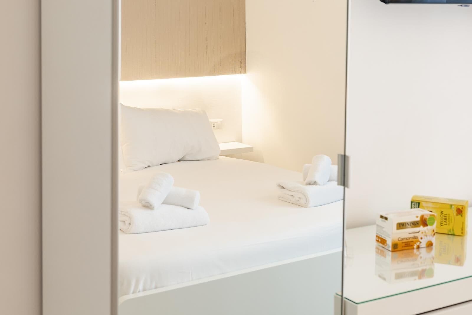 A hotel room with a bed, pillows, and neatly folded towels. There are two boxes of toiletries on the side table.
