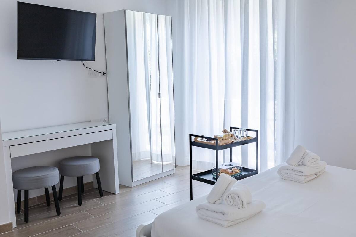 A hotel room with a bed, a desk with two chairs, a TV mounted on the wall, a breakfast trolley, and sheer curtains.