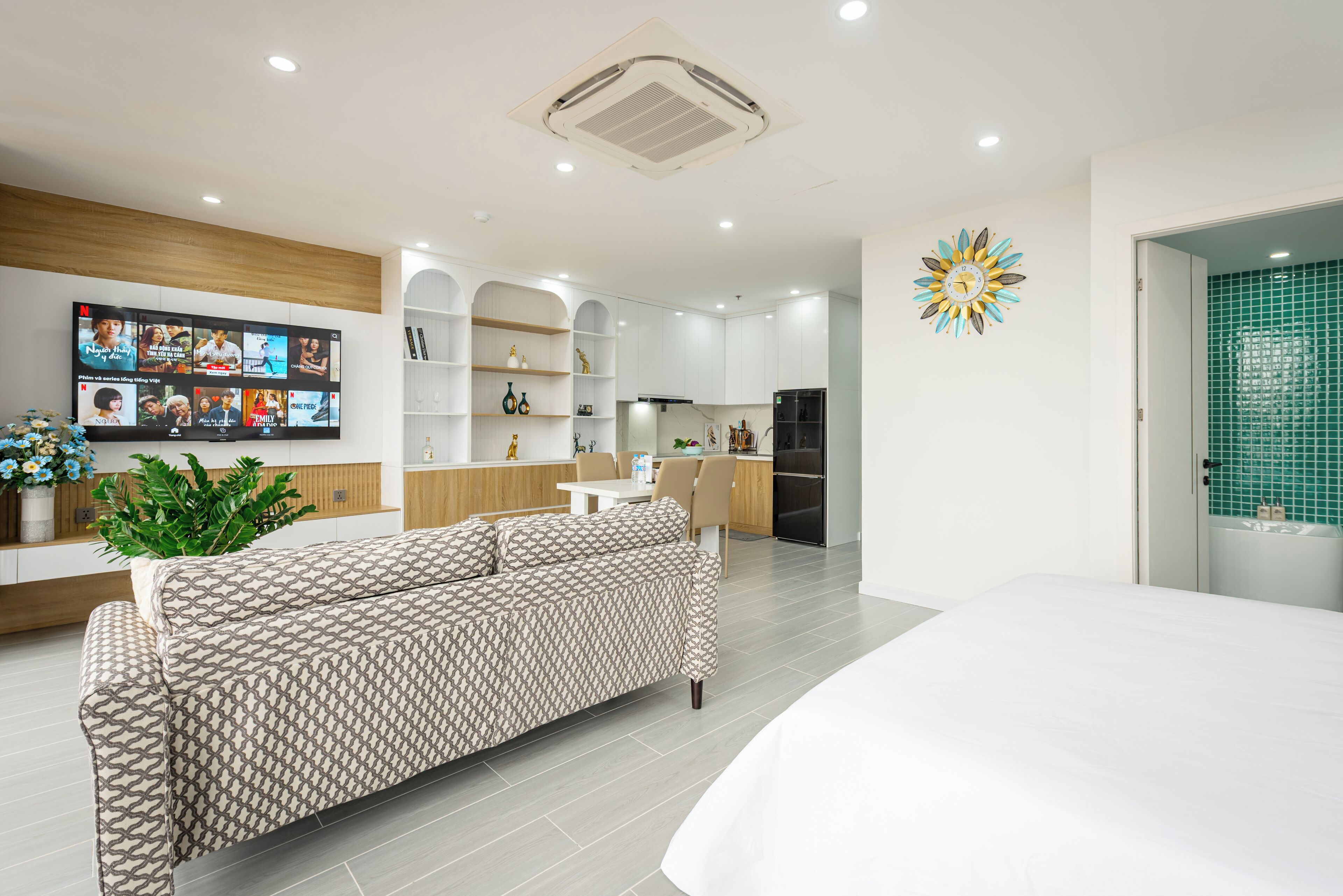 A modern living room with a sofa, a flat-screen TV, and a kitchen area with white cabinets and a refrigerator.