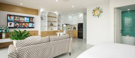 A modern living room with a sofa, a flat-screen TV, and a kitchen area with white cabinets and a refrigerator.