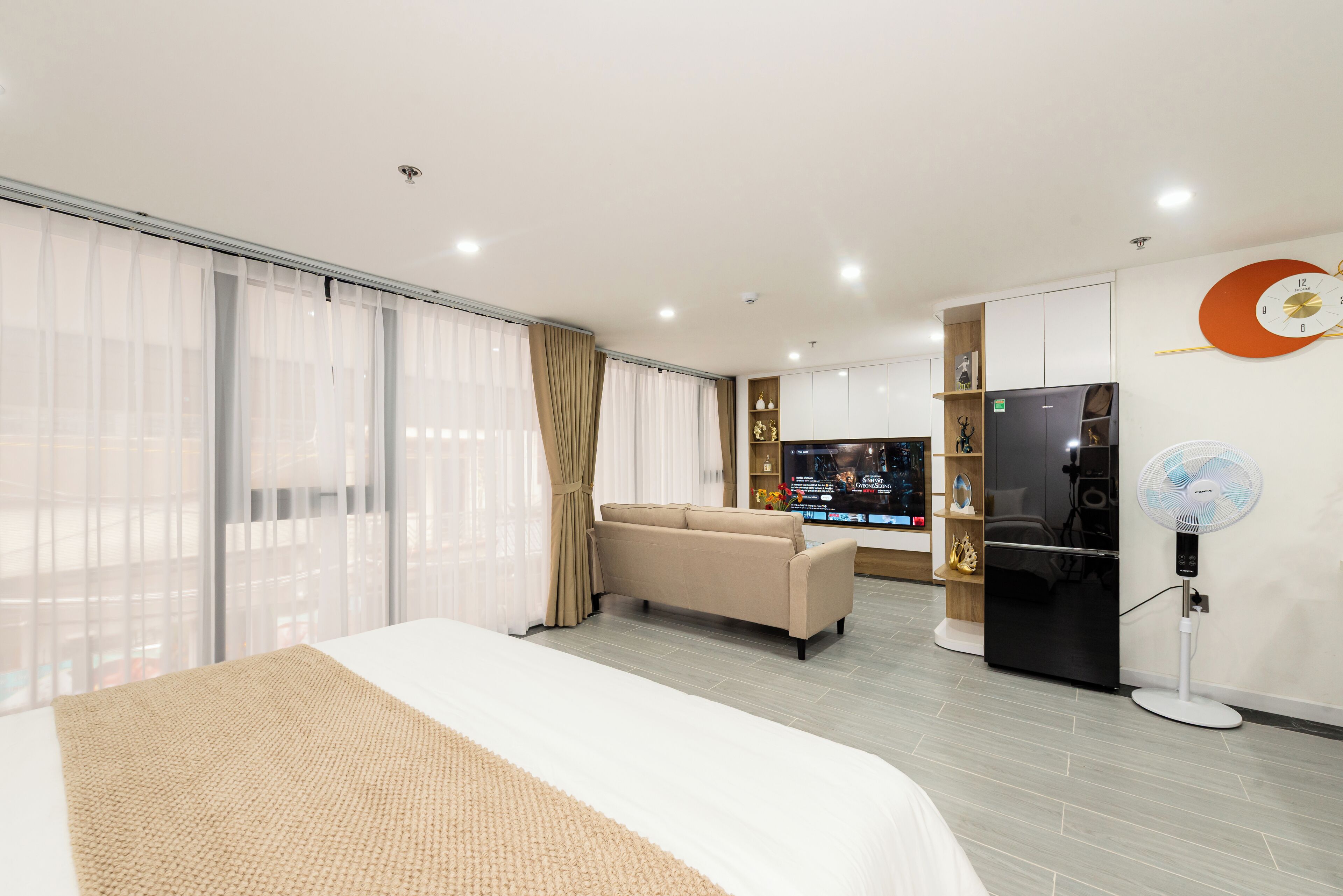 A modern hotel room with a large bed, a sofa, a refrigerator, a fan, and a TV in the background.