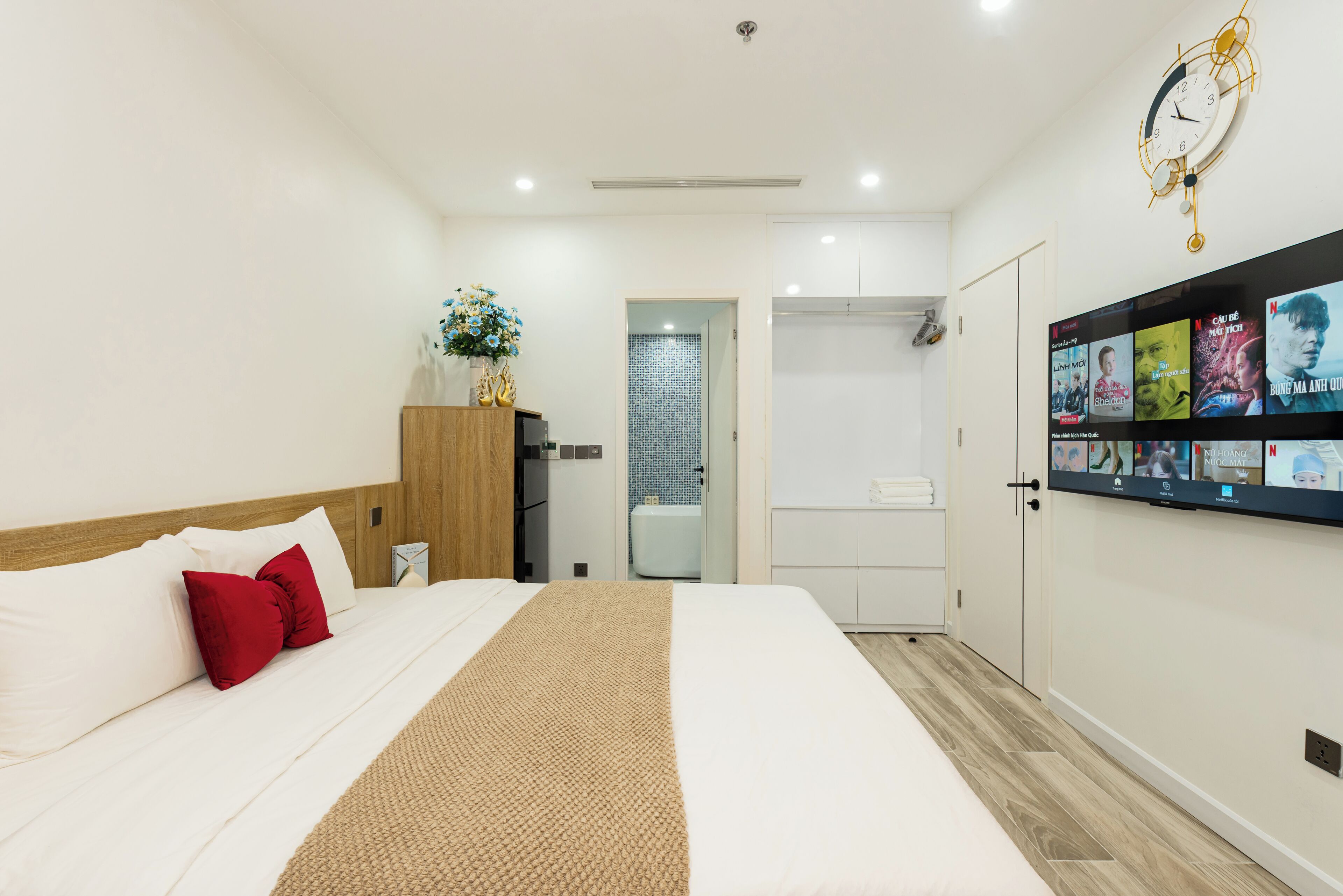 A modern hotel room with a large bed, a wooden headboard, a red pillow, a beige throw blanket, a wall-mounted TV, and a clock.
