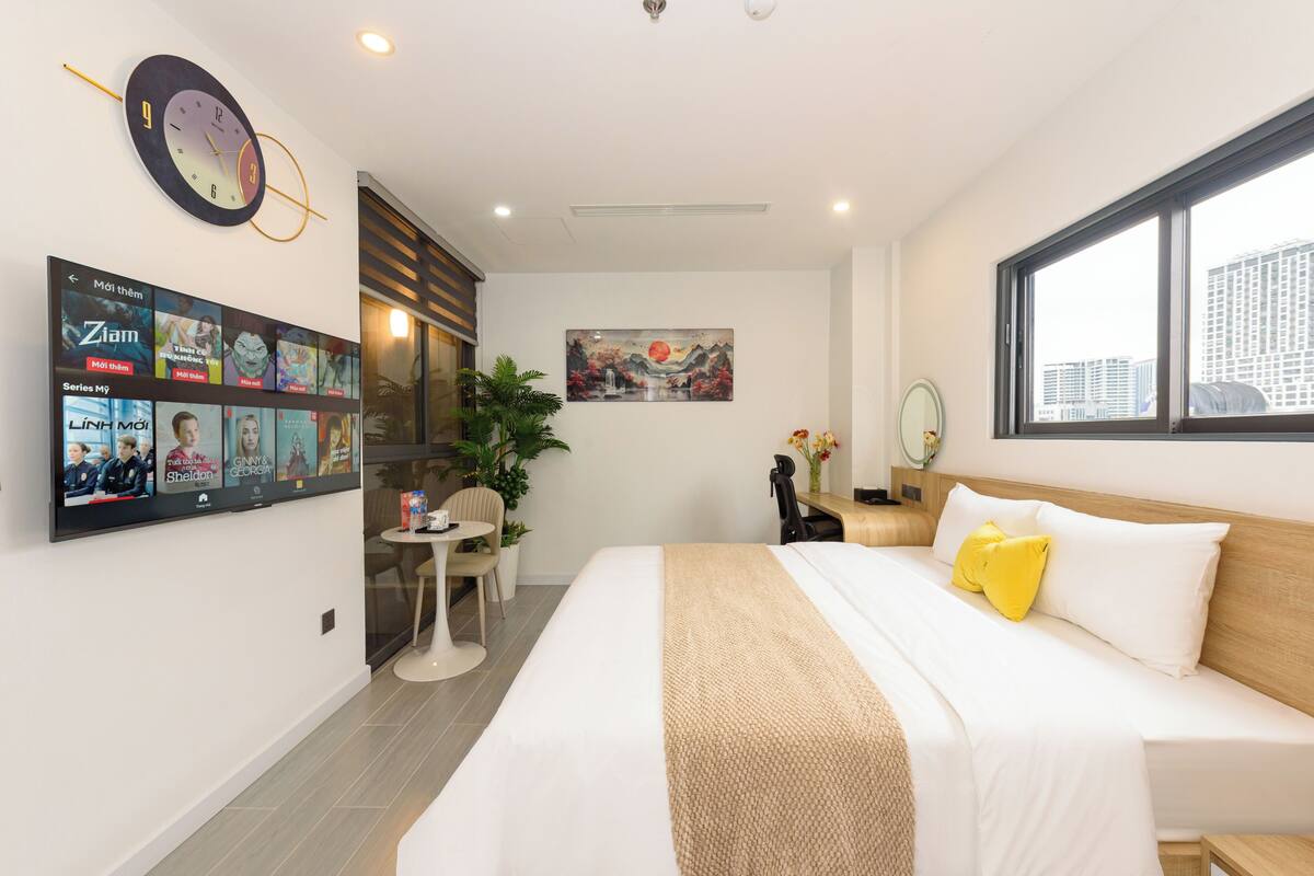 A modern hotel room with a large bed, a TV mounted on the wall displaying movie posters, a painting of a landscape, and a window with a city view.
