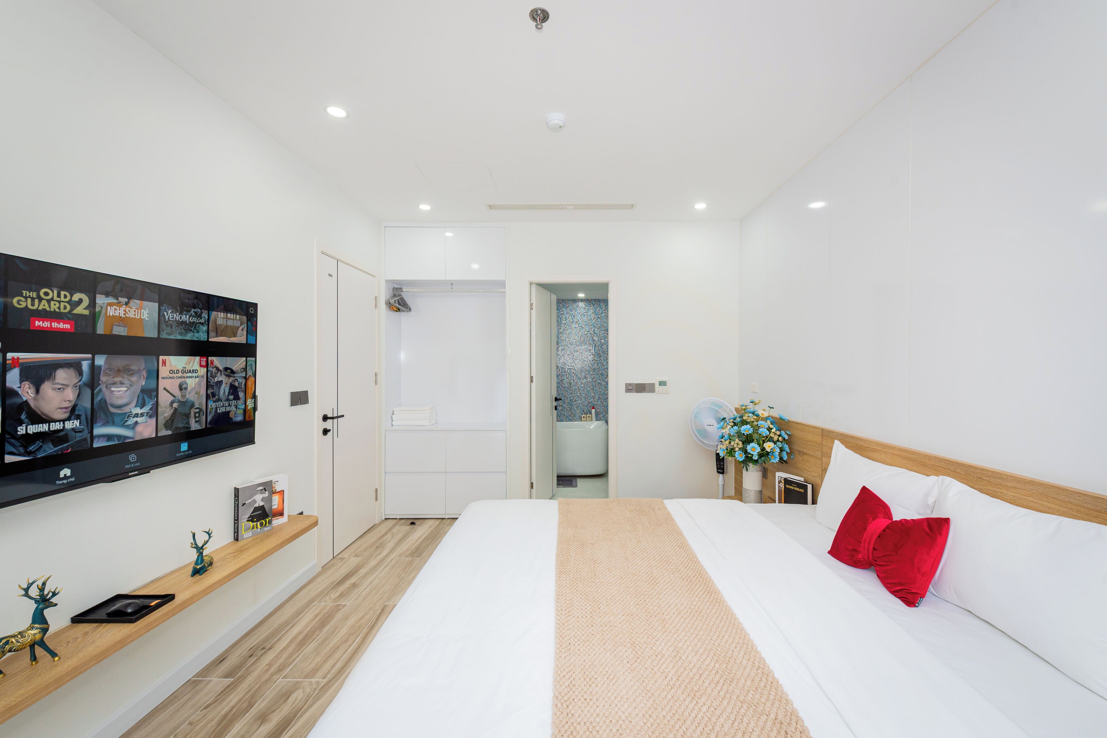 Ett modernt hotellrum med en stor säng, en tv som är monterad på väggen, en trähylla med dekorativa föremål och ett badrum som syns genom en öppen dörr.
