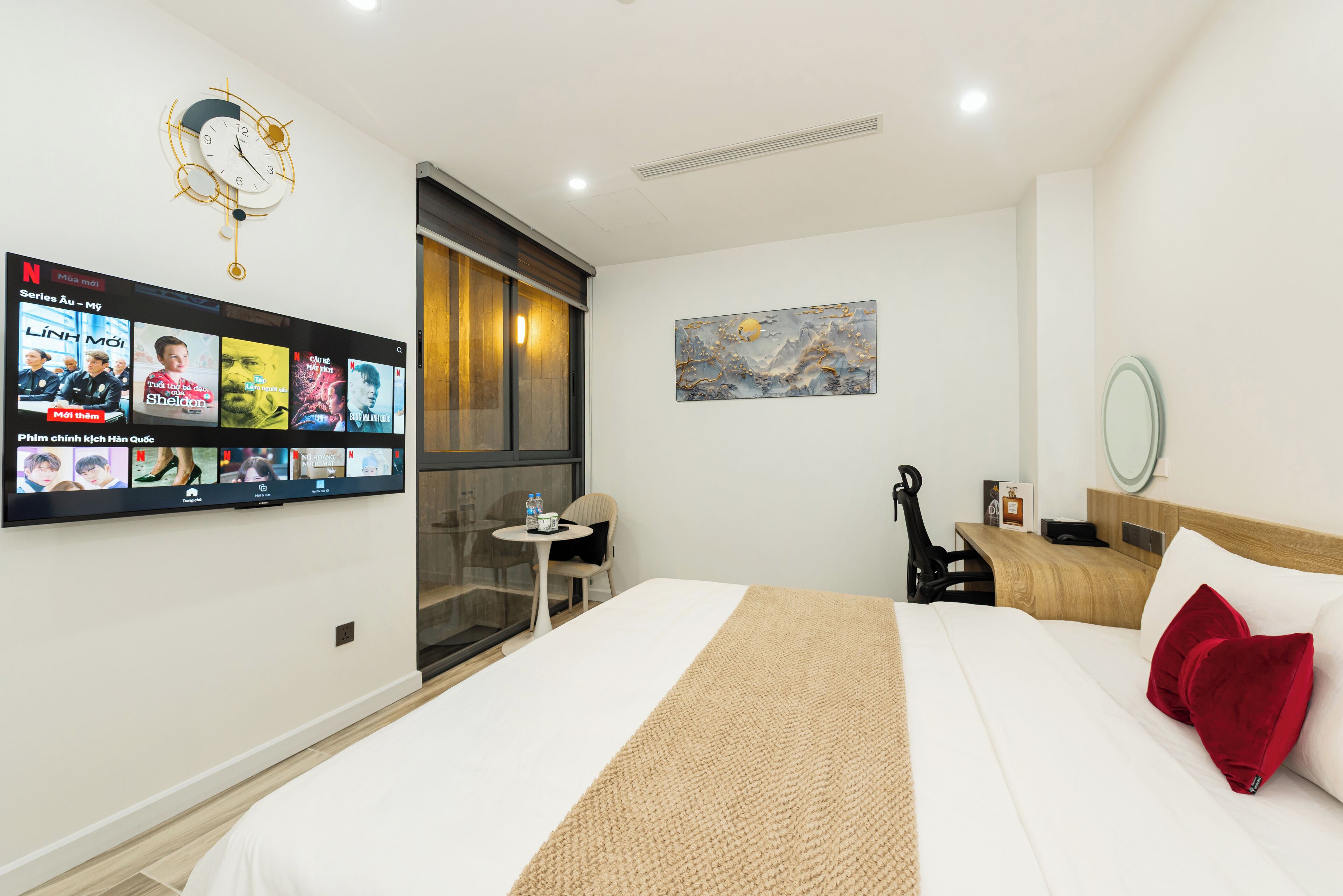 A hotel room with a bed, a TV mounted on the wall displaying Netflix content, a small table with a chair, and a painting on the wall.