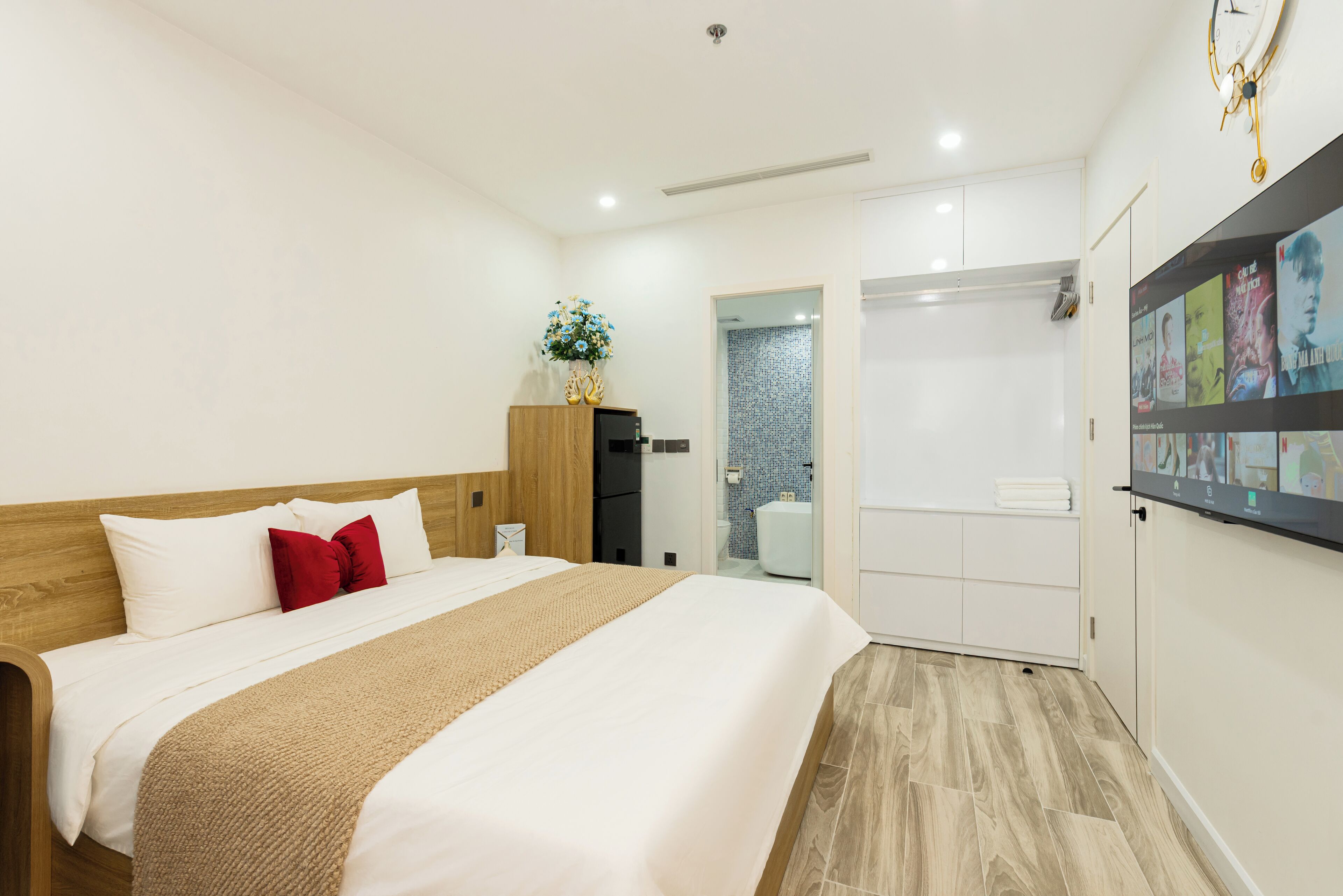 A modern hotel room with a wooden headboard, a large bed with white linens, a red pillow, and a beige throw blanket. There is a small wooden cabinet with a vase of flowers, a white door leading to a bathroom, and a wall-mounted shelf with books and a clock.