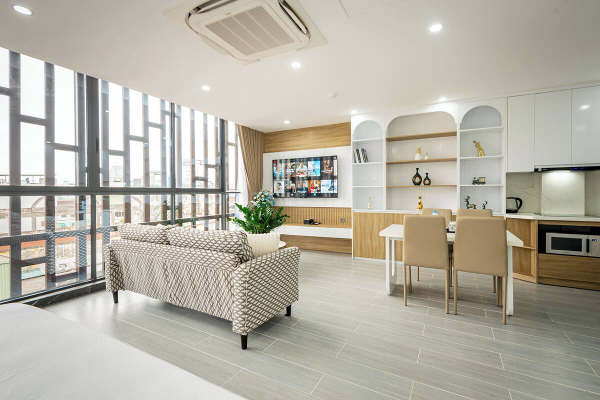 A modern living room with a sofa, a dining area with chairs, a kitchen with cabinets, and a large window with curtains.