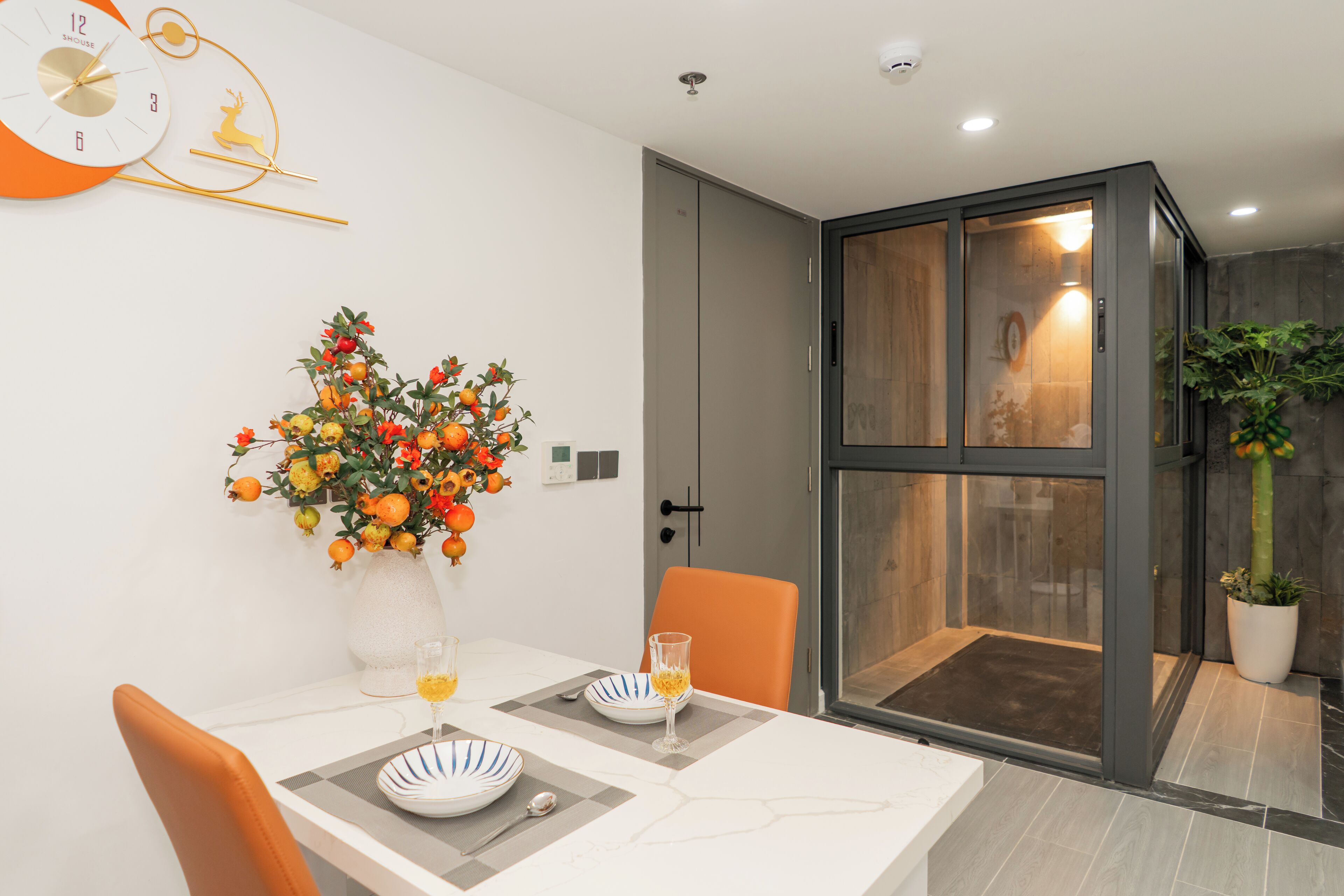 A modern dining area with a table set for two, a decorative orange and yellow flower arrangement, and a large window with a view of the outdoors.