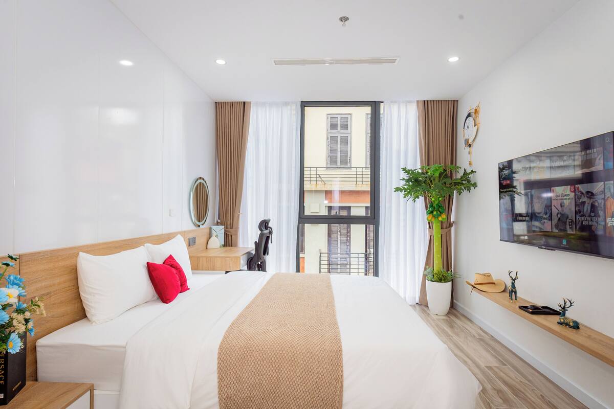 A modern hotel room with a large bed, a TV mounted on the wall, a potted plant, and a balcony view.