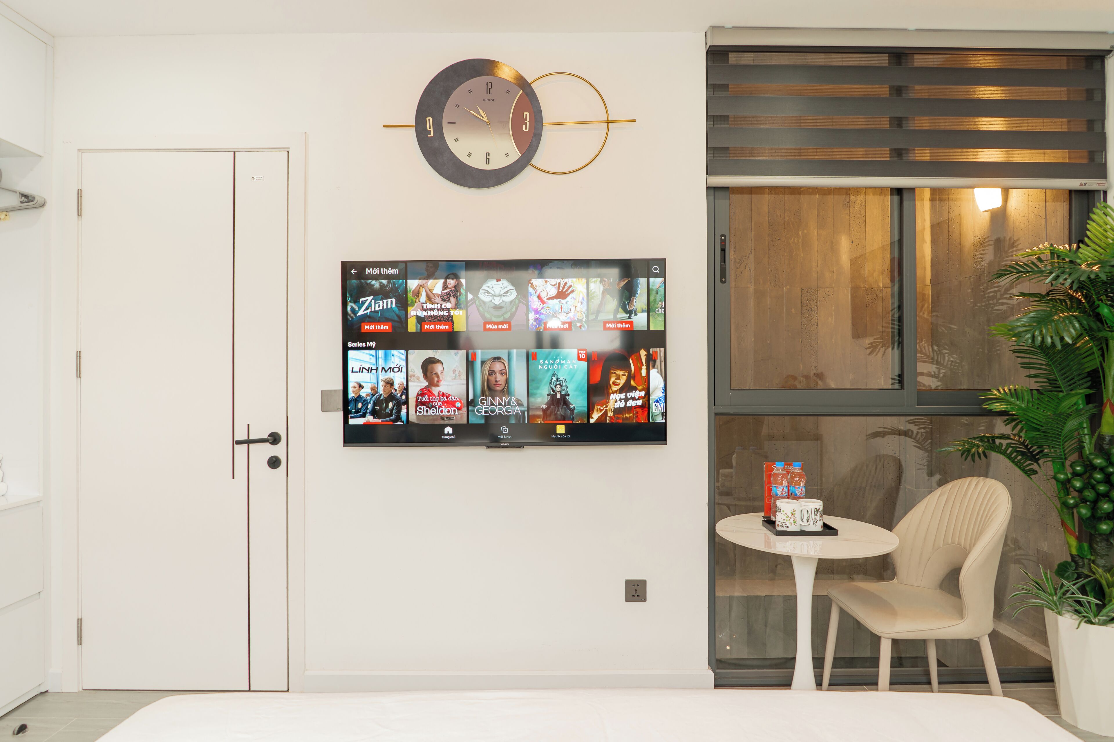 A modern living room with a flat-screen TV mounted on the wall displaying various shows, a round wall clock, a small round table with a chair, and a potted plant.