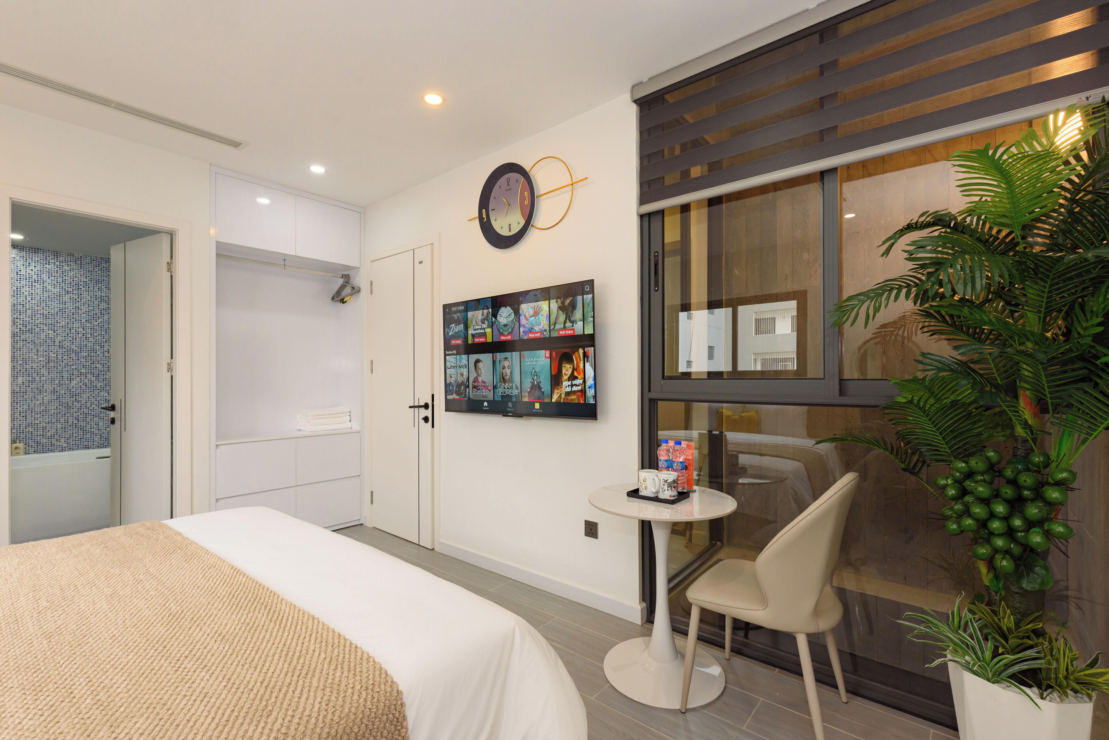 A modern hotel room with a bed, a small table with a chair, a TV mounted on the wall, and a large window with a view of the outside.
