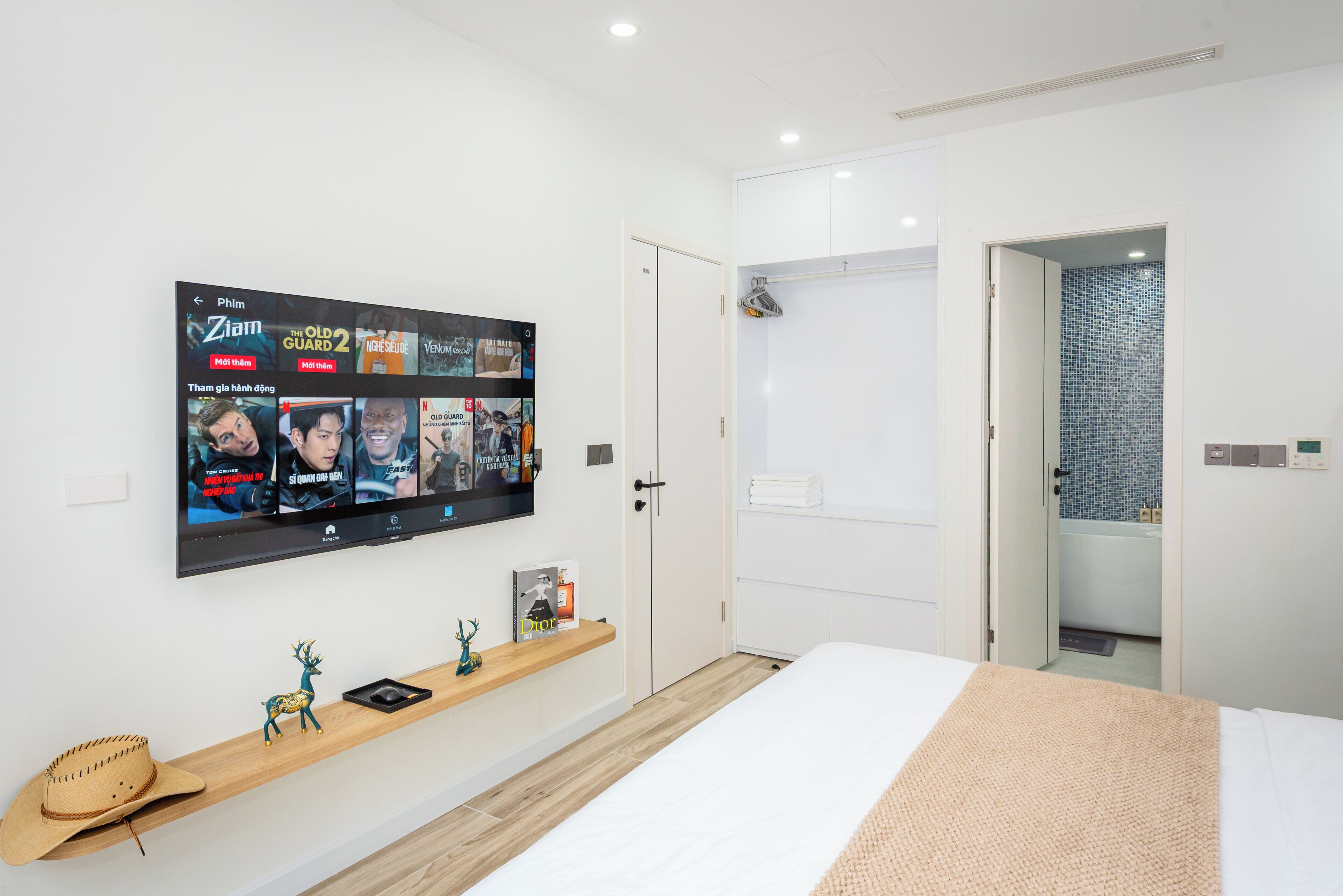 Ett modernt hotellrum med en platt-TV som är monterad på väggen och visar olika filmer. Det finns en hylla i trä med dekorativa föremål och en hatt. Rummet har en stor säng med ett beige täcke och en vit dörr som leder till ett badrum med en dusch som har blå kakelplattor.