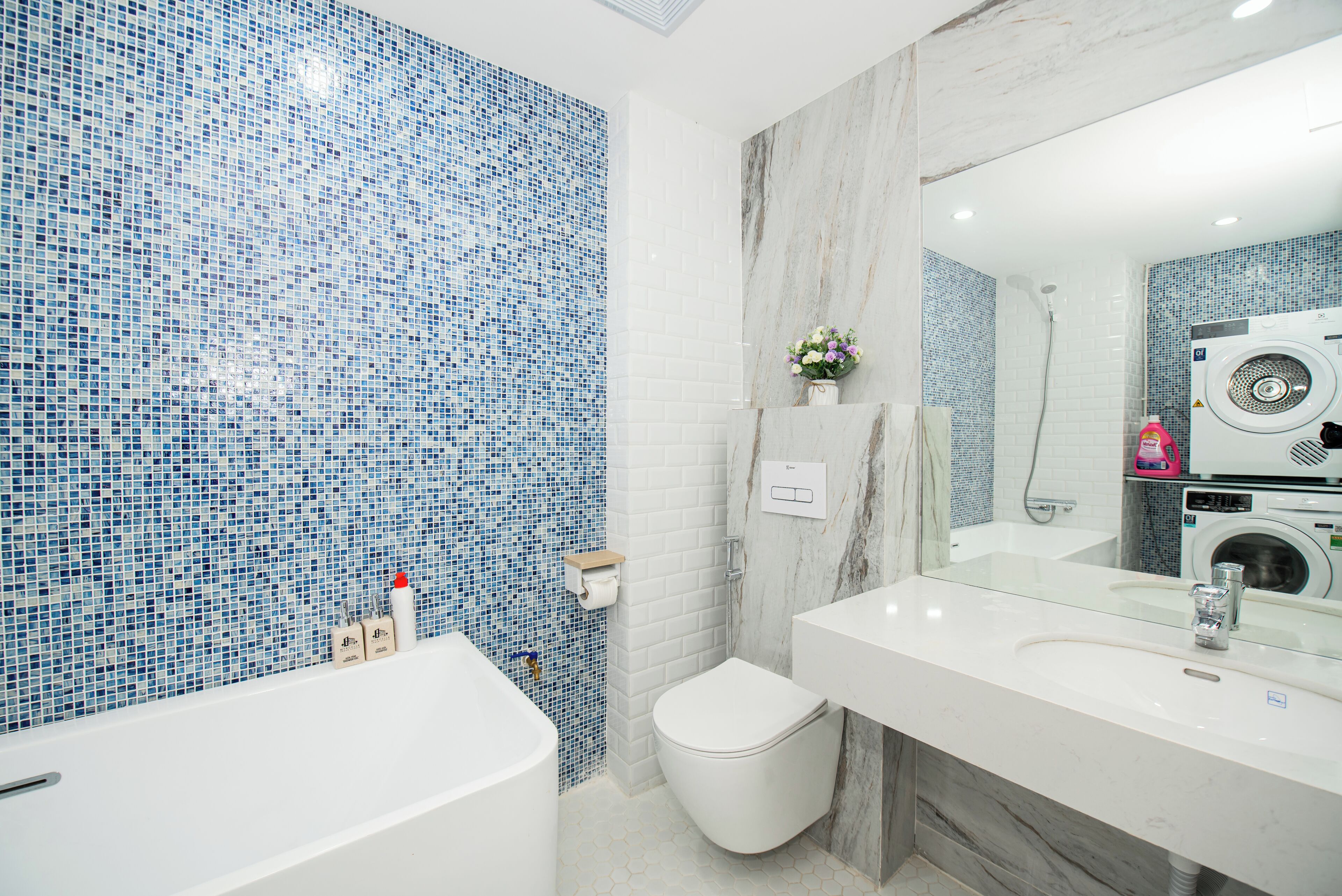 Une salle de bain avec un mur recouvert de mosaïques, des toilettes blanches, un lavabo avec un miroir et un lave-linge.