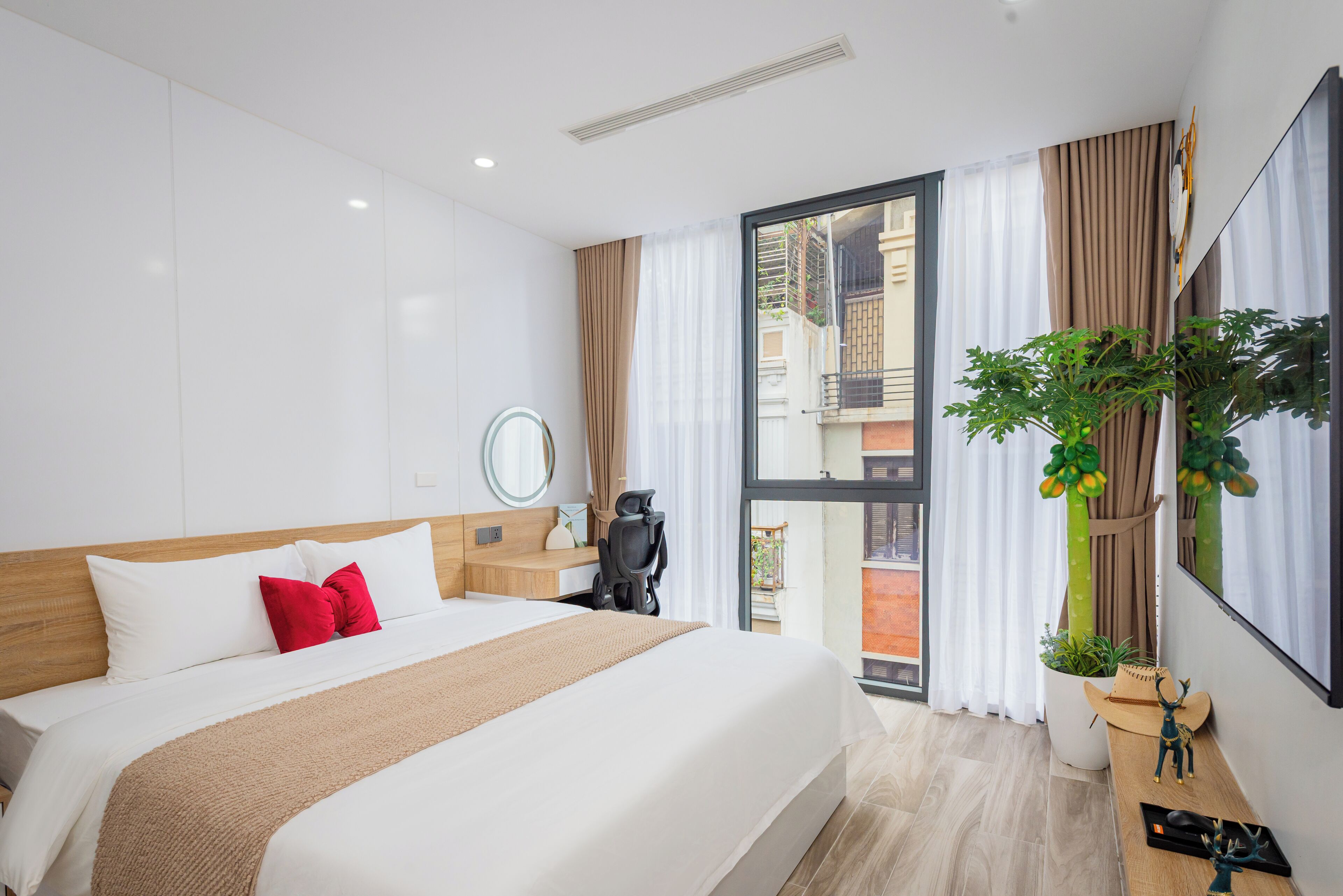 A modern hotel room with a large bed, a desk, a chair, a potted plant, and a view of the outside balcony.